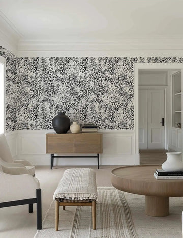 Lifestyle shot of a living room featuring animal print wallpaper with a repeating pattern of black leopard spots and speckles on a white background. The wallpaper covers the upper half of the walls, above white wainscoting. The room includes a light wood sideboard with black legs, a large black vase, a smaller textured cream vase, and books. Two cream armchairs are visible on the left, and a light wood coffee table with a white vase and books is on the right, all on a textured cream rug.