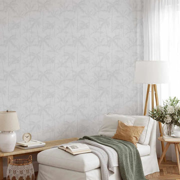 "Antigua wallpaper featuring a repeating pattern of hand-drawn palm trees in a soft, muted grey on a white background, creating a subtle tropical motif. The wallpaper covers a wall in a bright living room, behind a white chaise lounge with a green throw and a brown pillow. A wooden side table with a lamp and a basket are visible on the left, and a floor lamp and flowers are on the right."