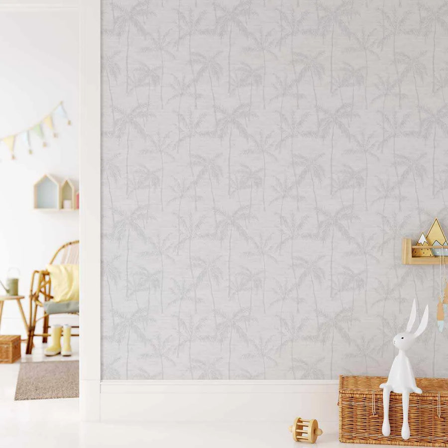 Antigua Wallpaper displayed on a nursery wall featuring a repeating pattern of tall palm trees in soft gray and beige tones on a light cream background. The design shows vertical palm fronds creating a tropical forest effect. The wallpaper is shown installed in a bright, minimalist child's room with natural wood furniture, wicker baskets, white decorative objects, and gold-toned accessories on shelves.