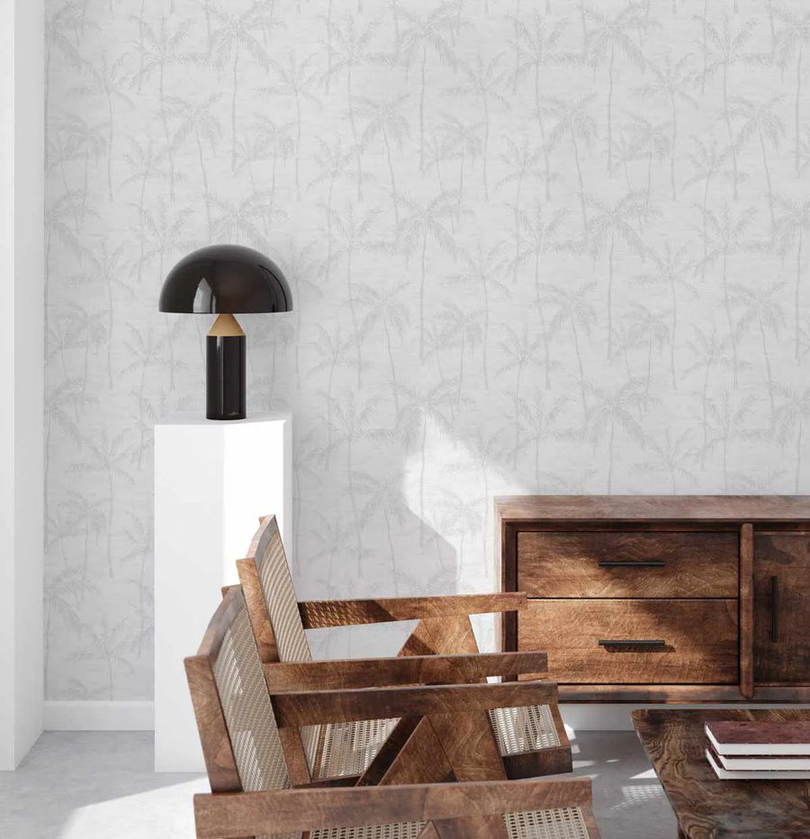 "Antigua Wallpaper featuring a repeating pattern of palm trees in a subtle, light grey tone on a white background, creating a soft, tropical texture on the wall of a living room. The room includes a black mushroom-shaped table lamp on a white pedestal, two wooden chairs with woven cane backs, and a dark wooden sideboard with drawers and doors."