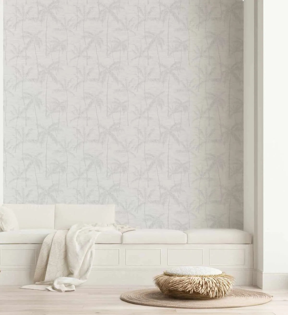 Antigua Wallpaper displayed on a bedroom wall featuring a repeating pattern of delicate palm fronds in soft gray and cream tones on a light cream background. The wallpaper has a subtle, understated botanical design with vertical paneling lines. Below, a cream-colored upholstered bed with matching bedding, a draped cream throw blanket, and a woven natural fiber pouf with fringe detailing on a circular jute rug complete the serene, minimalist room setting.