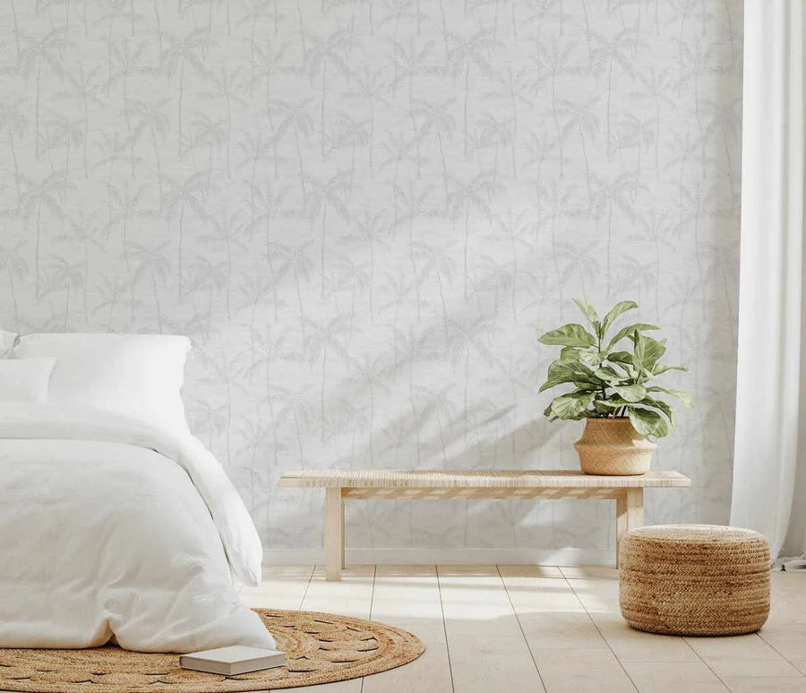 "Bedroom with a white bed, a wooden bench, and a potted plant, featuring Antigua Wallpaper on the back wall. The wallpaper has a repeating pattern of palm trees in a light grey tone on a white background, creating a subtle tropical feel. The room includes a round woven rug and a woven pouf, adding natural textures."