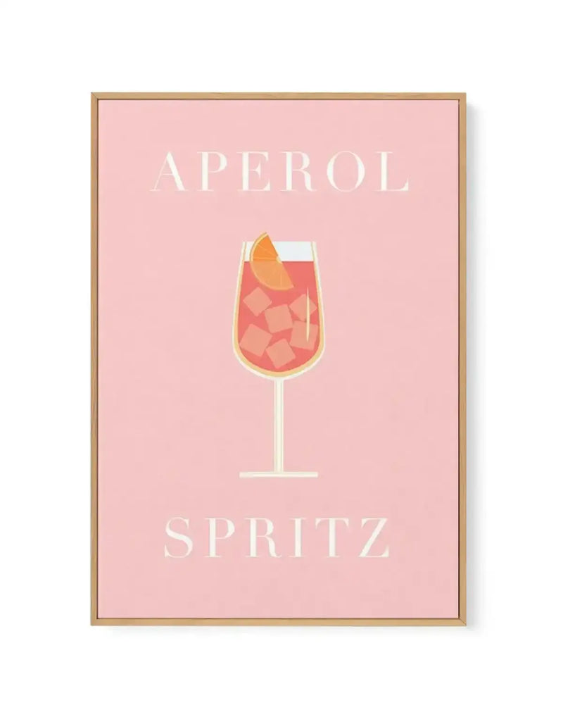 Framed canvas art print featuring the word "APEROL" in white capital letters at the top and "SPRITZ" at the bottom, centered on a soft blush pink background. The focal point is a minimalist illustration of an Aperol Spritz cocktail in a wine glass with a golden rim, filled with coral-red liquid and ice cubes, garnished with an orange slice. The artwork has a clean, modern aesthetic with a warm natural wood frame and white border.