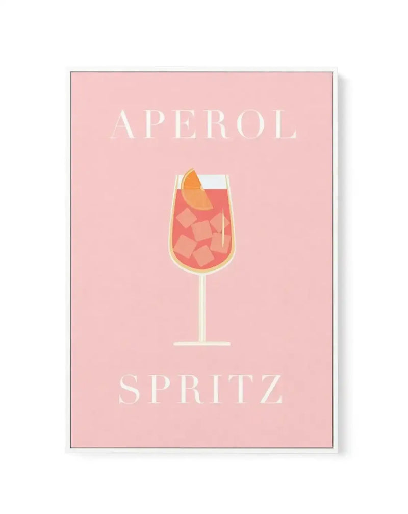 Framed canvas art print featuring a minimalist illustration of an Aperol Spritz cocktail on a dusty rose pink background. The drink illustration shows a wine glass filled with coral-red liquid and ice cubes, garnished with an orange slice, rendered in warm peachy and coral tones with a cream-colored glass outline. The text "APEROL" appears at the top and "SPRITZ" at the bottom in elegant cream-colored capital letters. The canvas has a gray frame with white border.