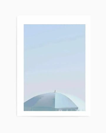 "Photography art print featuring the top of a light blue beach parasol against a clear, pale blue sky. The parasol is seen from a low angle, showing its curved shape and the small white finial at its peak. The image has a soft, minimalist aesthetic with subtle variations in the blue tones of the parasol and sky."