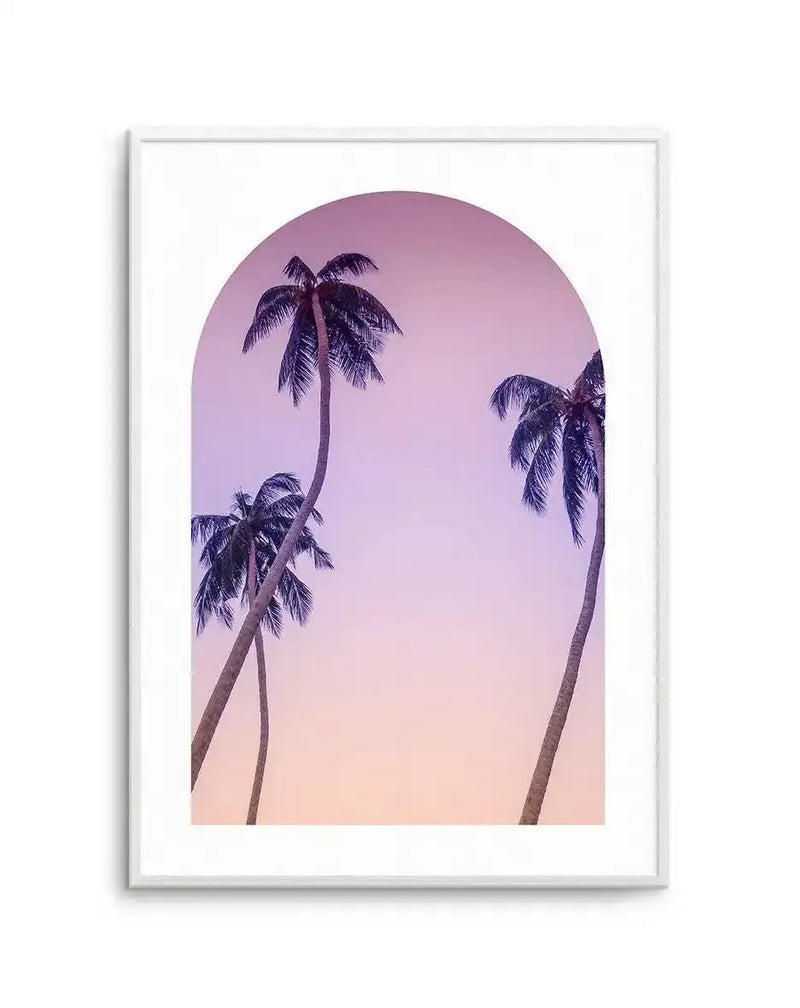 Photography art print featuring the silhouettes of three palm trees against a gradient sky, transitioning from lilac purple at the top to soft peach at the bottom, framed within a white arched border and displayed in a white frame with a white border.