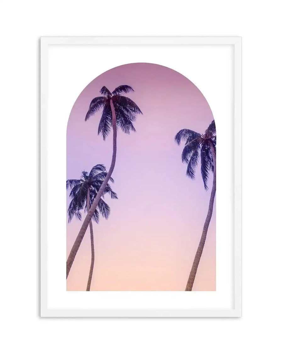 Framed art print featuring a semi-circular archway revealing a tropical scene with three palm trees silhouetted against a gradient sky. The sky transitions from soft lavender at the top to a warm peach hue at the bottom. The palm trees have dark fronds and slender trunks, with two on the left curving towards the center and one on the right curving inwards. The print is displayed in a white frame with a white border.
