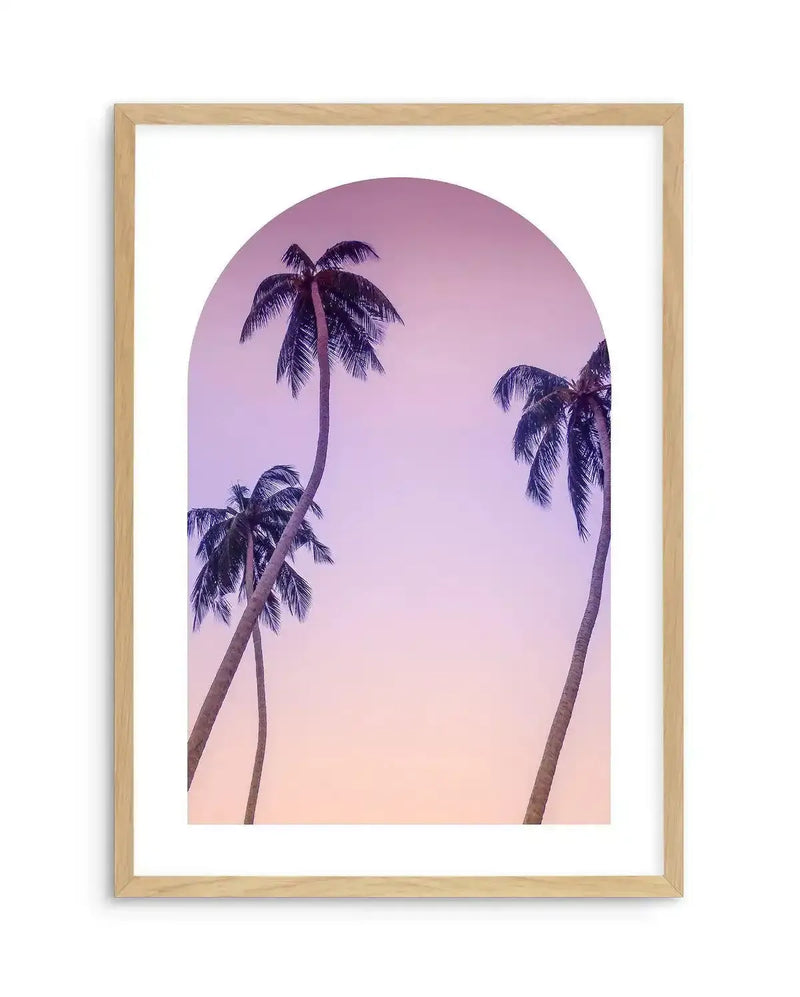 Framed photography art print with a natural wood frame and white border, featuring three palm trees silhouetted against a gradient sky. The sky transitions from a soft lavender-pink at the top to a warm peach-orange at the bottom, all contained within an arched window shape.
