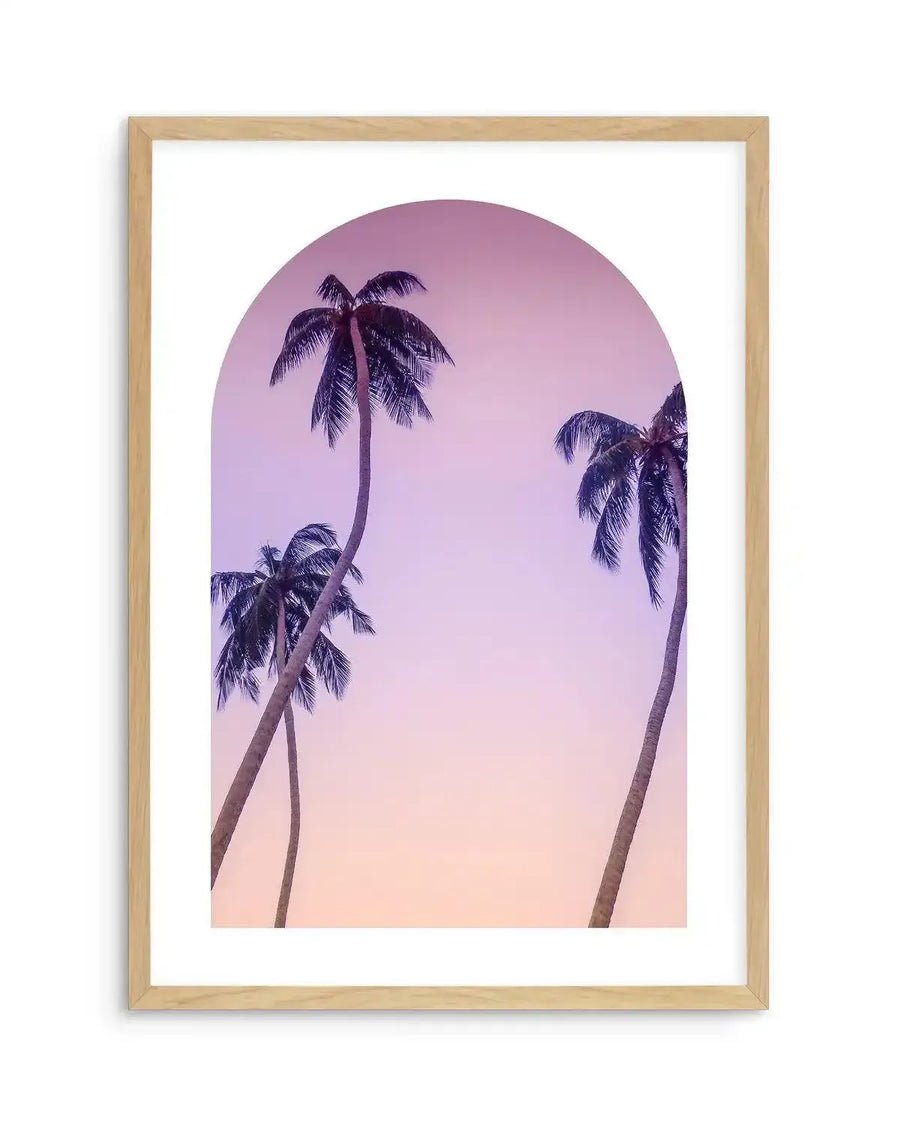 Framed photography art print with a natural wood frame and white border, featuring three palm trees silhouetted against a gradient sky. The sky transitions from a soft lavender-pink at the top to a warm peach-orange at the bottom, all contained within an arched window shape.