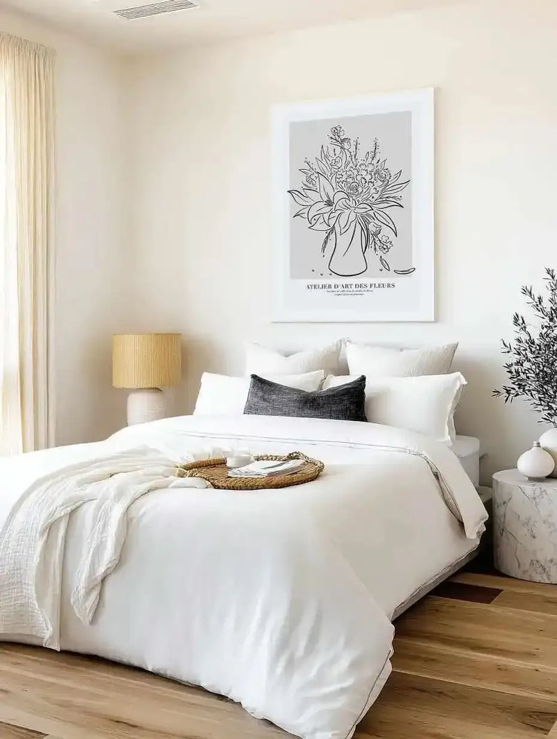 Large framed art print displayed above a bed in a minimalist bedroom. The print features a black and white line drawing of a bouquet of flowers in a vase on a light grey background, with a white frame and white border. The room includes a bed with white bedding, a dark grey throw pillow, a woven tray, a bedside lamp with a woven shade, and a marble side table with a plant.
