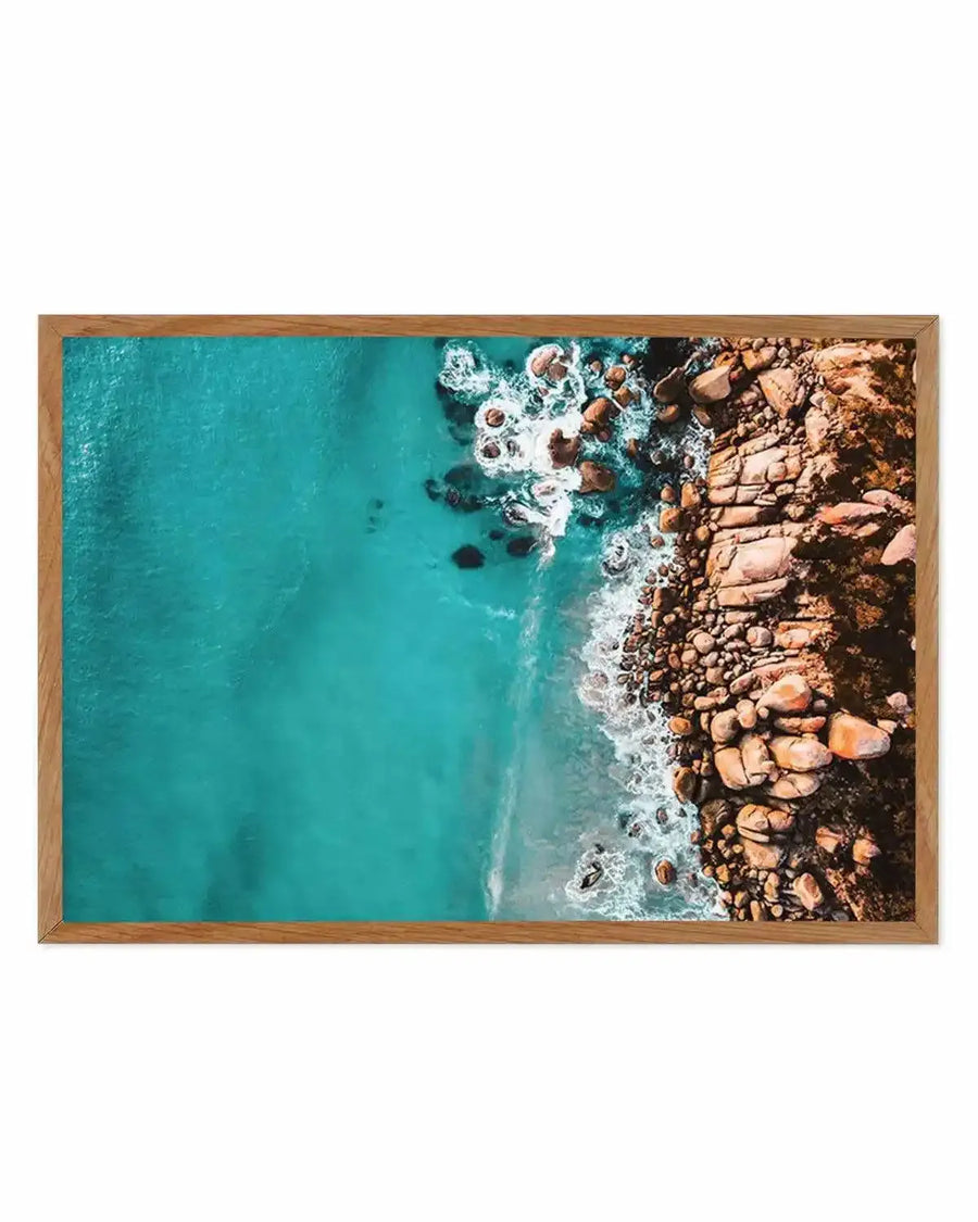 Framed aerial photography art print of a rugged coastline with a natural oak frame and white border. The composition features vibrant turquoise ocean water on the left, meeting a rocky shore on the right where waves crash and foam, revealing dark brown and orange-hued boulders.