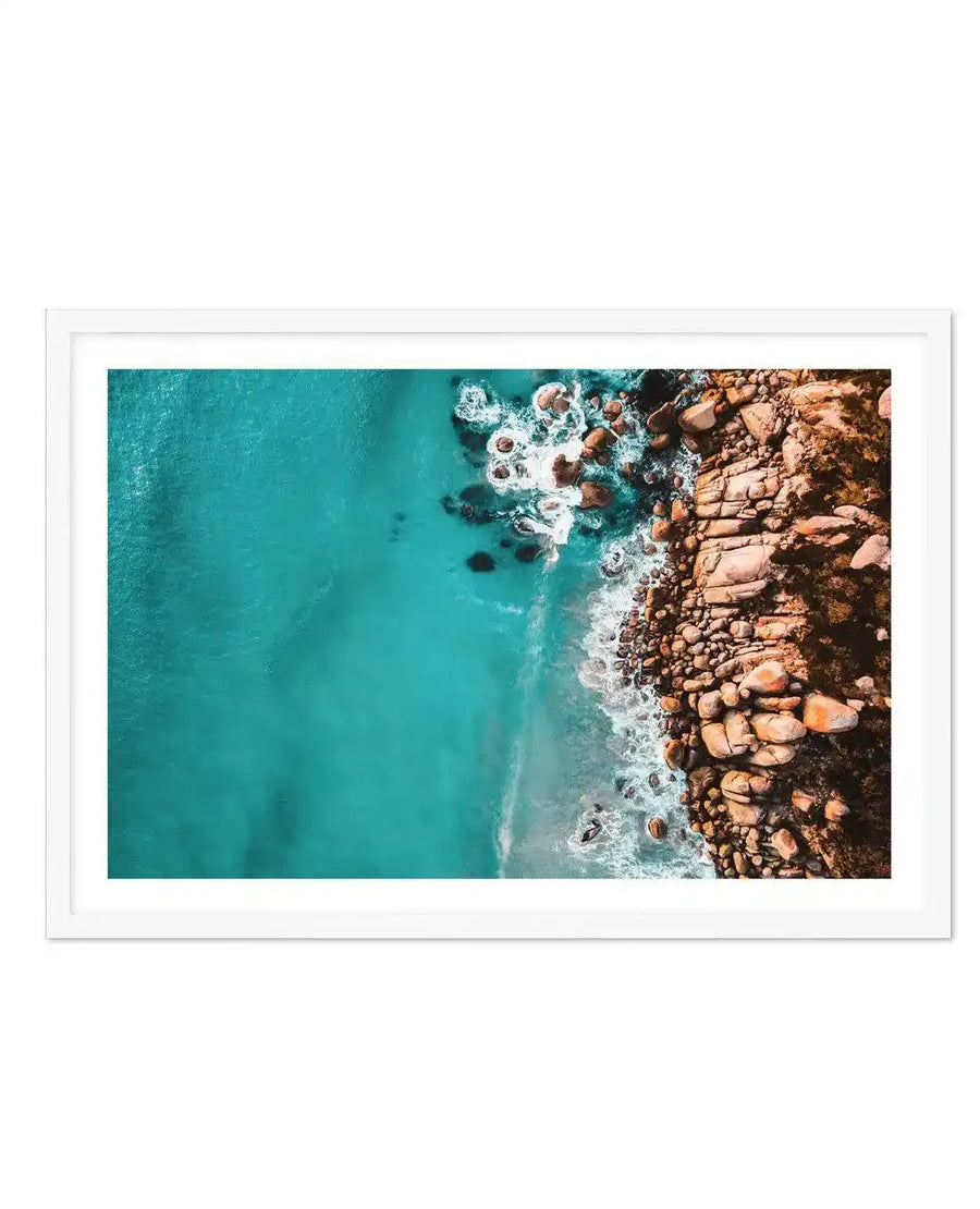 Framed aerial photograph of a vibrant turquoise ocean meeting a rocky coastline, with a white frame and white border. The composition captures the clear blue-green water on the left, transitioning to a sandy shore and large, warm-toned boulders on the right, with white waves crashing against the rocks.