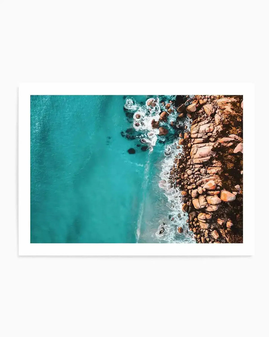 Aerial photography art print of a rocky coastline meeting a turquoise ocean. The image shows the deep blue-green water on the left, with white waves crashing against a sandy shore and a rugged, reddish-brown rocky outcrop on the right. The rocks vary in size and shape, creating a textured landscape where they meet the foamy white water.
