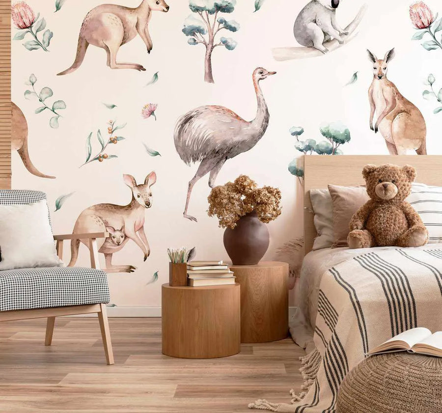 Peel and stick wallpaper featuring a repeating pattern of hand-painted Australian wildlife and flora in soft watercolour tones on a pale cream background, displayed in a child's bedroom. The design includes a koala, kangaroos, an emu, and various native plants in shades of muted green, dusty pink, and warm brown. The wallpaper is visible on the wall behind a bed with a light wood headboard, a striped throw blanket, a teddy bear, and a small armchair, creating a serene and naturalistic atmosphere.