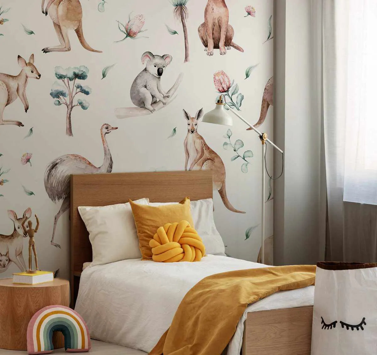 "Australian wildlife wallpaper displayed in a child's bedroom above a wooden bed with white bedding and mustard yellow throw and cushions. The wallpaper features hand-painted watercolour illustrations of various Australian animals including kangaroos, a koala, and an emu, along with native plants like banksia flowers and eucalyptus leaves, on a light cream background. The room also includes a round wooden side table with a rainbow cushion and a white floor lamp."
