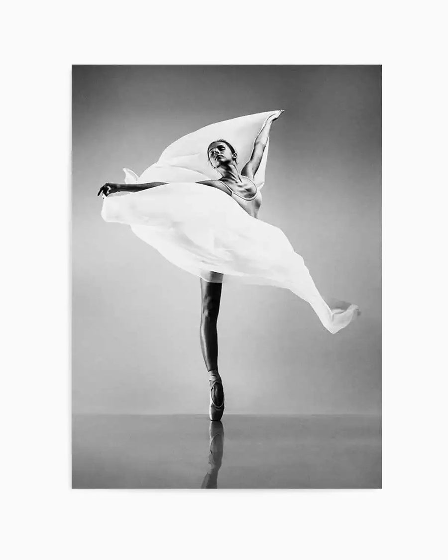 Framed black and white photograph of a ballerina en pointe, balancing on one foot with a large piece of flowing white fabric draped around her. Her arms are extended, one reaching up and the other forward, creating a dynamic and elegant pose. The dancer's reflection is visible on the polished floor, with a plain grey background.