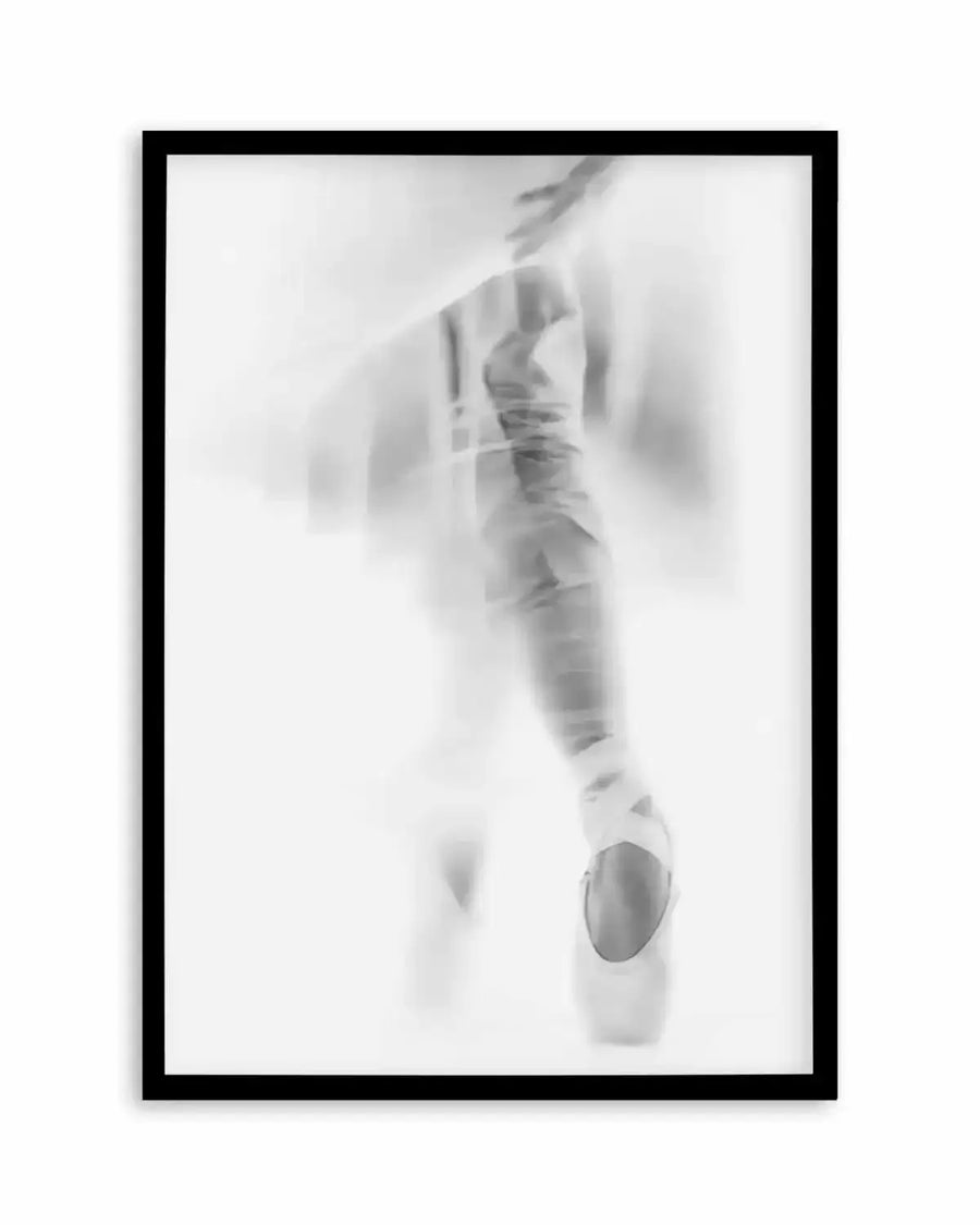 Framed black and white photograph of a ballerina en pointe with a black frame and white border. The composition captures the lower half of the dancer's body in motion, with a soft blur effect, creating a dreamlike and ethereal mood.