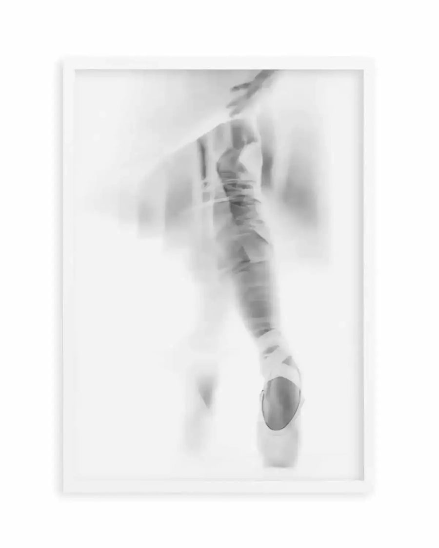 Framed black and white photograph of a ballerina en pointe, captured with a soft focus and motion blur effect, creating a dreamlike and ethereal composition. The ballerina's leg is extended, with her foot in a pointe shoe clearly visible, while her upper body and arms are softly blurred, suggesting movement. The image has a high-key aesthetic with bright whites and subtle grey tones, displayed in a white frame with a white border.