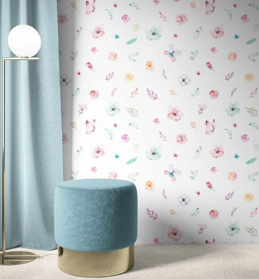 Peel and stick wallpaper featuring a repeating pattern of hand-painted watercolour flowers, leaves, and butterflies in soft pastel shades of pink, blue, green, yellow, and purple on a white background, displayed in a child's room. The room includes a light blue velvet ottoman with a gold base, a gold floor lamp with a spherical white light, and a light blue curtain.