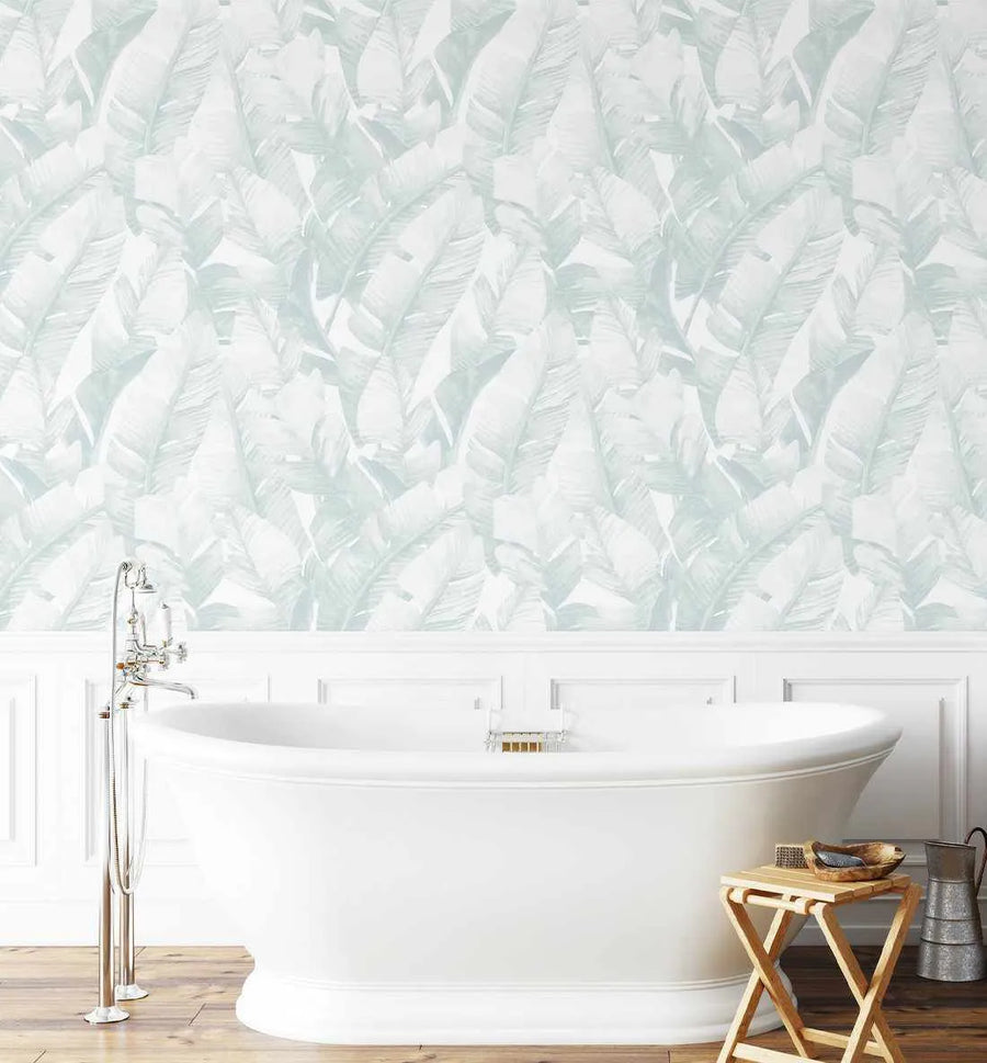 Banana Leaf Palms wallpaper in seafoam green and white displayed on a bathroom wall above white wainscoting. The design features an overlapping pattern of tropical banana leaves and palm fronds in soft sage green and cream tones with a watercolor illustration style. The wallpaper is shown in a spa-like bathroom setting with a white freestanding soaking tub, brass fixtures, wooden flooring, and natural wood accessories, creating a serene, botanical atmosphere.