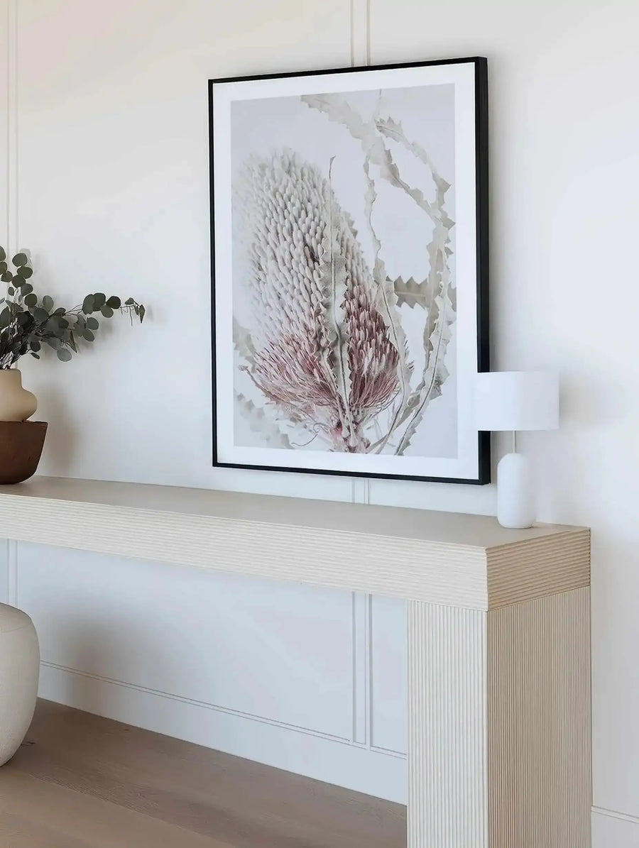 Lifestyle shot of a minimalist entryway with a framed art print displayed above a light wood console table. The print features a close-up photograph of a dried Banksia flower in muted tones of cream and dusty pink, with a black frame and white border. The console table has a fluted texture and is adorned with a small white lamp and a vase of eucalyptus branches.