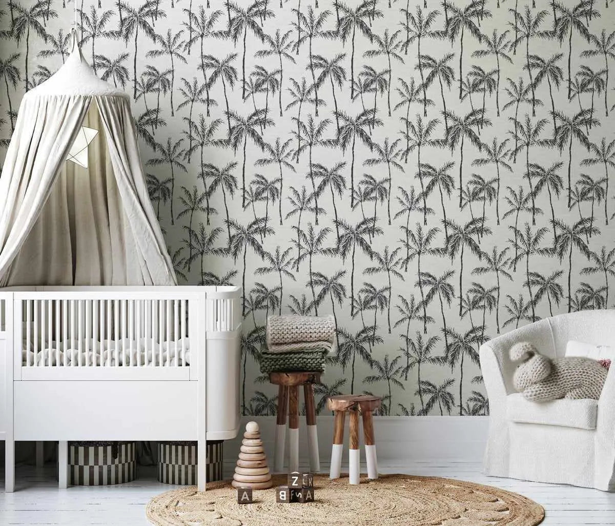 Nursery:
Nursery room with white walls featuring Barbados wallpaper with a repeating pattern of hand-drawn palm trees in black on a light cream background. A white crib with a light grey canopy is on the left, and a white armchair with a knitted toy is on the right. A round jute rug, wooden stools, and children's toys are on the floor.