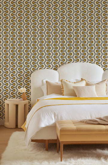 Lifestyle shot of a bedroom featuring Bardot Retro Floral Wallpaper. The wallpaper has a repeating pattern of stylized floral motifs in mustard yellow, black, and cream, creating a retro, geometric design. The wallpaper covers the wall behind a white, textured headboard and bed with white bedding and yellow accents. A light-colored, sculptural side table with a small vase of flowers is visible to the left of the bed, and a yellow upholstered bench sits at the foot of the bed on a white fluffy rug.