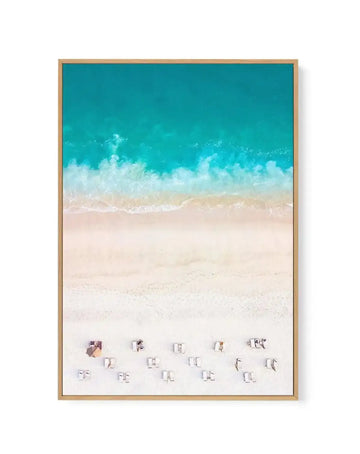 Framed canvas art print featuring an aerial beach scene with turquoise ocean water at top, white-capped waves in the middle, and pale sand below dotted with approximately fifteen beach umbrellas and lounge chairs scattered across the shore. The photograph has a soft, dreamy quality with warm beige sand tones contrasting against vibrant teal water. Natural wood frame with light tan finish surrounds the canvas.