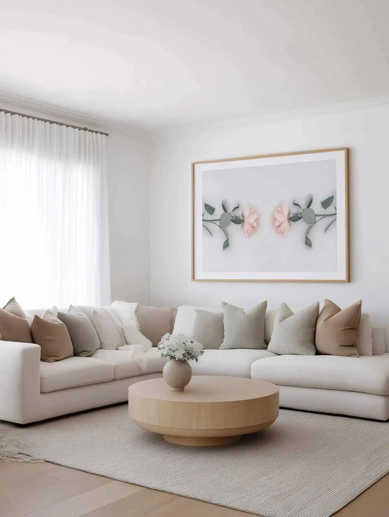 Lifestyle shot of a minimalist living room featuring a large framed art print of two pink roses on a white background, displayed above a curved white sectional sofa. The print has a natural wood frame and white border. The room includes a light wood round coffee table with a vase of white flowers, a textured cream rug, and sheer white curtains.
