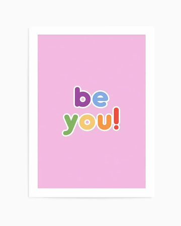 Art print featuring the words "be you!" in a playful, colourful font on a soft pink background, displayed in a white frame. The word "be" is in purple and light blue, and "you!" is in green, orange, and red, with a subtle white outline around each letter.