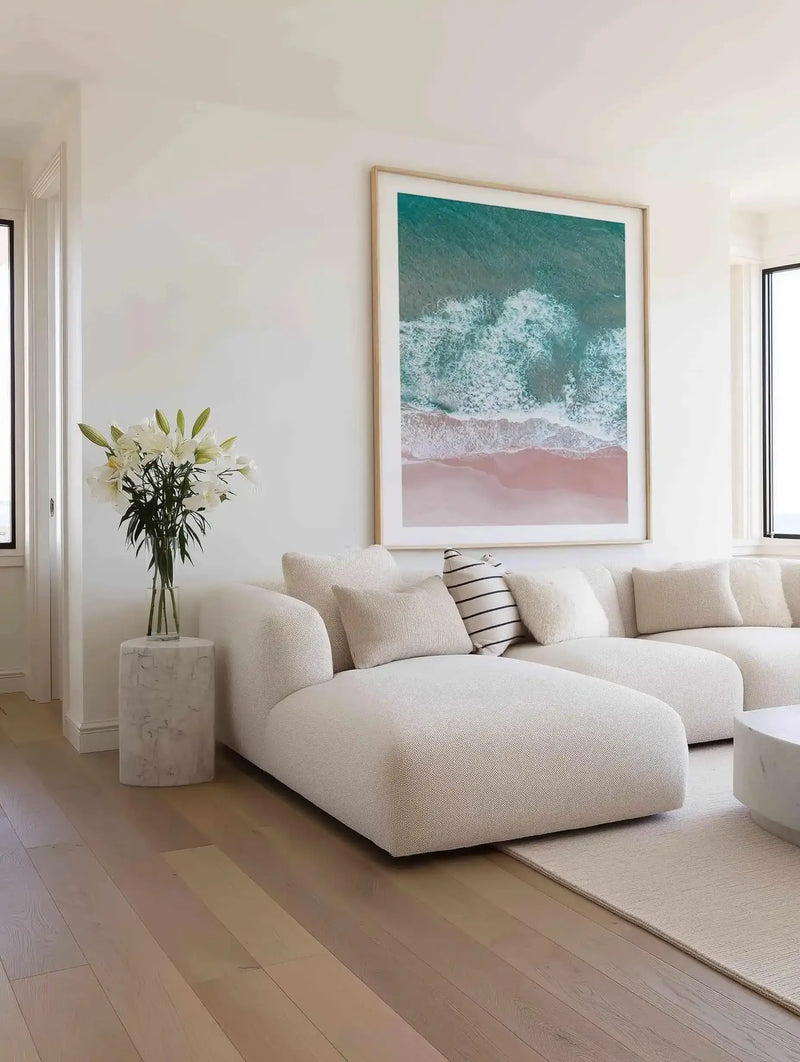 Aerial photograph art print titled "Beach Days" displayed in a natural wood frame with white border, showing turquoise ocean waves with white foam meeting a dusty rose-pink sandy beach. The print hangs above a curved cream-colored modular sofa with textured upholstery and coordinating throw pillows in a bright, minimalist living room with light wood flooring and white walls.