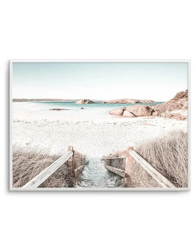 Framed photography art print of a serene coastal beach scene with a white frame and white border. The composition features a sandy beach in the foreground with a wooden fence leading into shallow, clear water. In the midground, the turquoise ocean stretches to the horizon with several large, smooth rocks and distant landmasses under a pale blue sky. The overall mood is calm and natural, with muted tones of sandy beige, soft blue, and light brown.