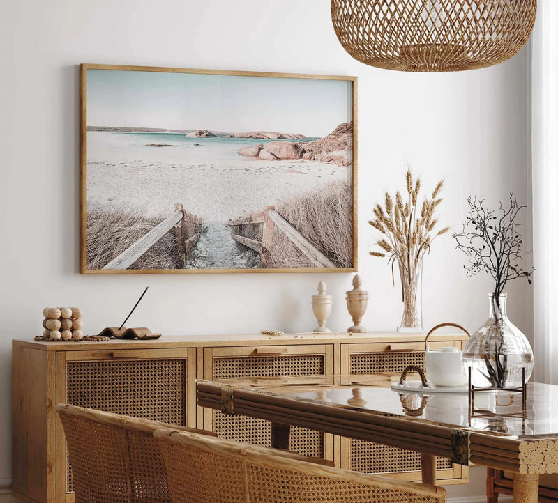Lifestyle shot of a dining room featuring a large framed art print above a wooden buffet. The print is a photograph of a sandy beach path leading to the ocean, with dry grass on either side and large rocks in the distance under a pale blue sky. The print is in a natural wood frame with a white border. The room includes a woven rattan pendant light, dried wheat stalks in a clear vase, and a glass-topped dining table with wooden chairs.