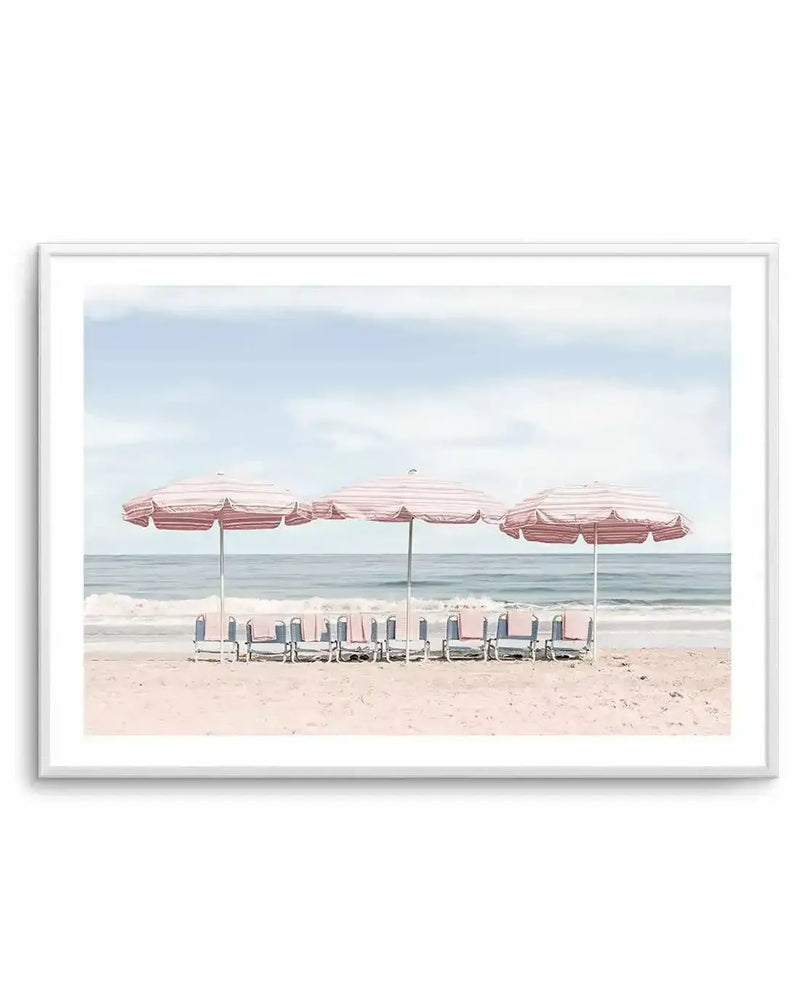 Framed photography art print of a beach scene with three pink and white striped beach umbrellas and matching chairs lined up on light pink sand, facing the calm blue ocean under a pale blue sky with soft white clouds, displayed in a white frame with a white border.