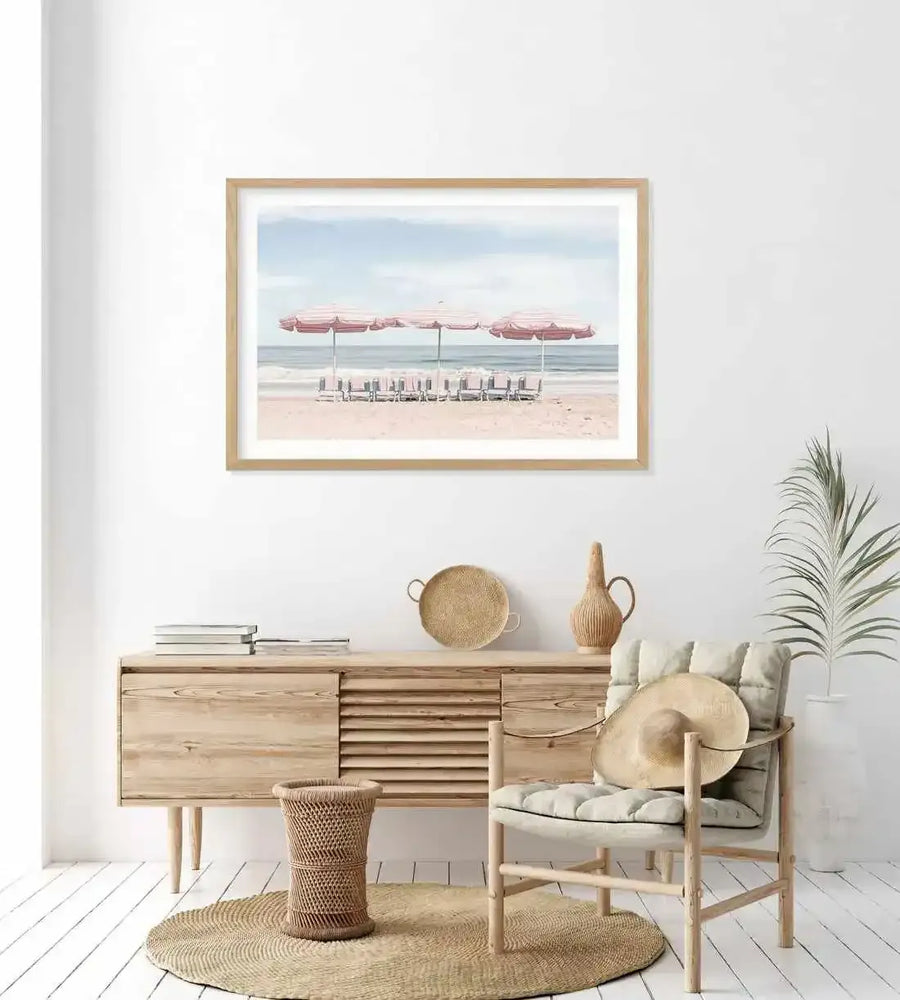 Lifestyle shot of a minimalist living room featuring a large framed art print of a beach scene with pink umbrellas. The print is displayed above a light wood sideboard. The artwork depicts three striped pink and white beach umbrellas with matching lounge chairs on a sandy beach, with a calm blue ocean and pale blue sky in the background. The room includes a woven rug, a wicker basket, a light wood armchair with a straw hat, and a potted plant.