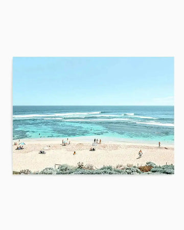 Aerial photograph of a pristine beach scene featuring pale golden sand dotted with beachgoers and colorful umbrellas, turquoise shallow waters with white-capped waves, and a clear light blue sky. Native coastal vegetation with silvery-green foliage borders the foreground. The print is unframed and displays the photograph with a clean white border on all sides, mounted on a neutral background.