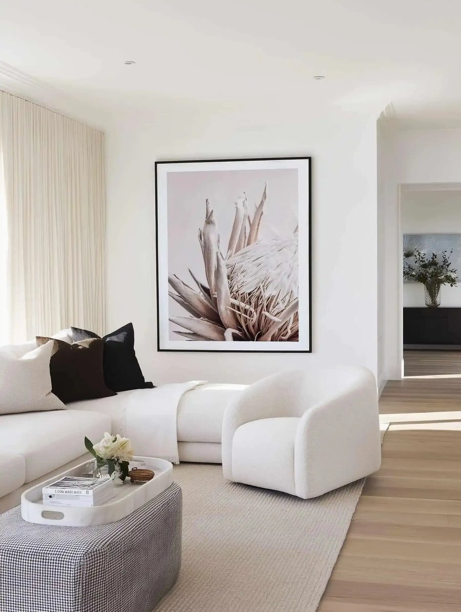 Beige Protea art print with black frame displayed on a white wall above a white curved chair. The print features a close-up photograph of a protea flower with dusty rose and taupe petals against a soft, pale pink background, shown with a white border. The room features minimalist décor including cream curtains, a white sectional sofa with black pillows, a grey textured ottoman with white tray, and light wood flooring.