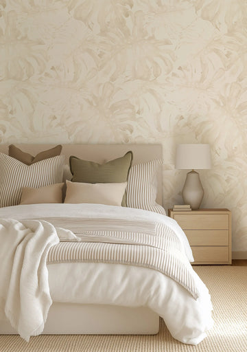 "Bedroom featuring a bed with a beige headboard and neutral bedding, with a bedside table and lamp. The wall behind the bed is covered in wallpaper featuring a repeating pattern of large-scale monstera leaves in a muted beige and cream watercolour style. The leaves have a soft, organic quality with subtle texture."
