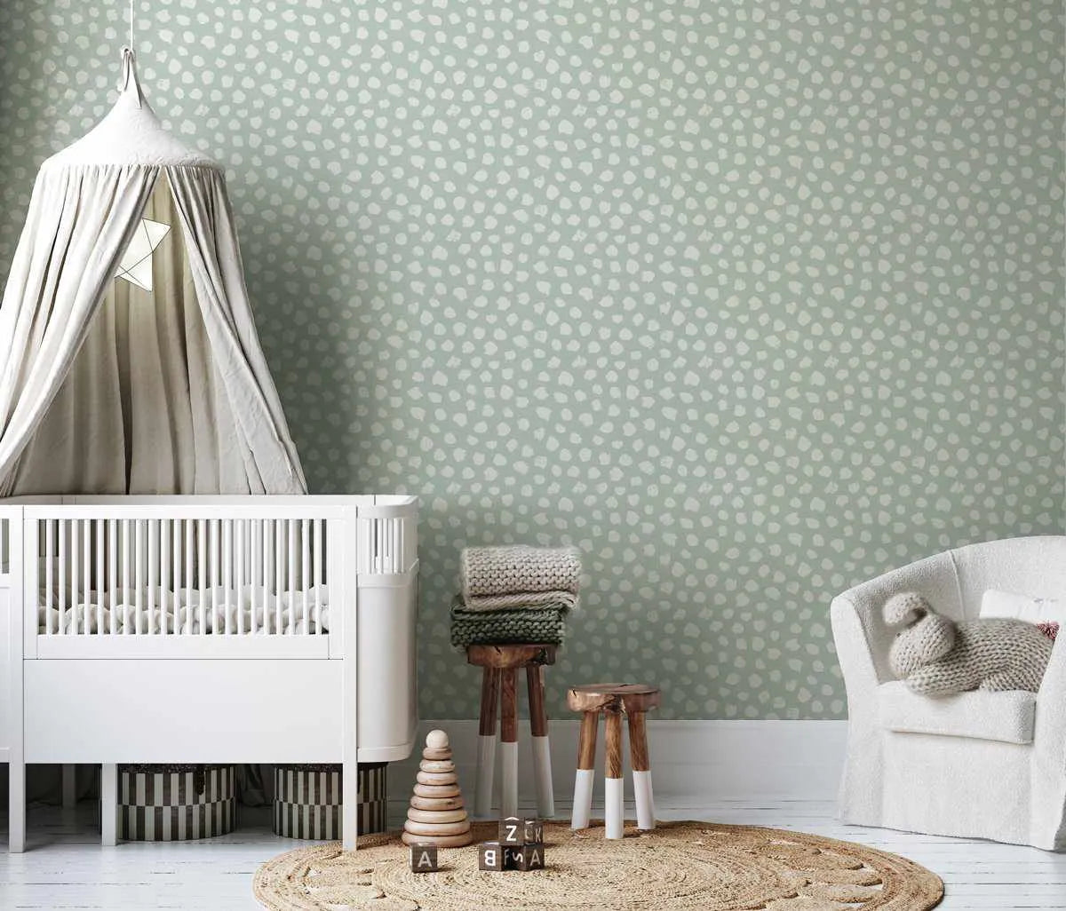 Bella's Dots Modern Mint Wallpaper displayed on a nursery room wall behind a white crib with canopy, white armchair, and wooden stools. The wallpaper features a repeating pattern of small white polka dots scattered across a soft sage green background. The design has a subtle, matte finish with even dot spacing creating a calming, minimalist aesthetic suitable for children's spaces.