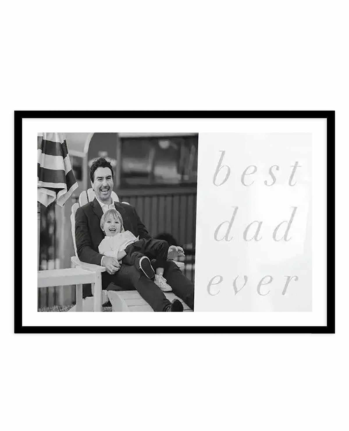 Black-framed art print featuring a black and white photograph on the left side showing a smiling man holding a young child while sitting outdoors with a striped umbrella visible behind them. The right side displays "best dad ever" in light gray, handwritten-style script text. White border surrounds the composition within the frame.