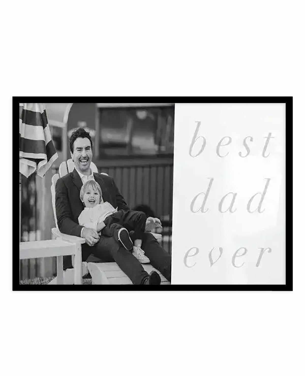 Black and white art print featuring a photograph on the left side showing a smiling man holding a young child on his lap, both seated outdoors with a striped flag visible in the background. The right side displays light gray serif text reading "best dad ever" in an elegant, handwritten style. The design is mounted in a black frame with white border, creating a horizontal landscape composition suitable for wall display.