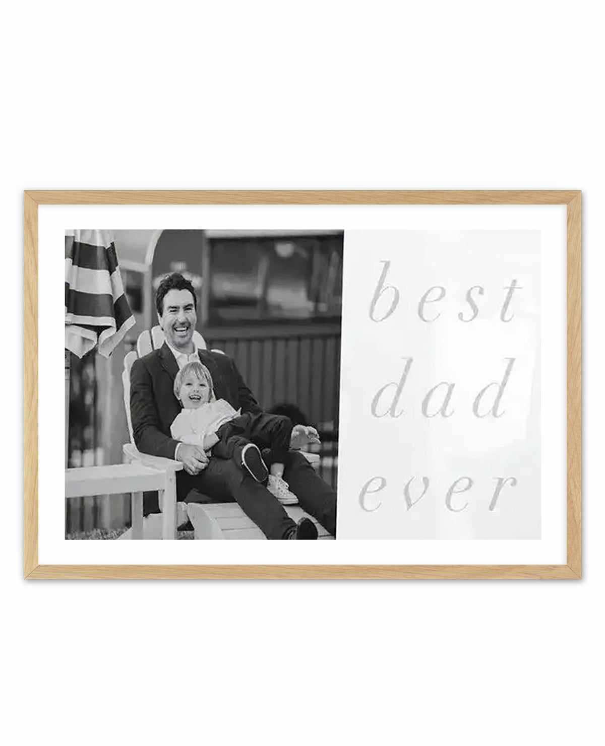 Custom art print in natural wood frame with white border featuring a black and white photograph of a man and young child sitting together on the left side, with light gray cursive text reading "best dad ever" on the right side. The photograph shows warm, candid emotion with an American flag visible in the background. The print has a clean, minimalist design combining photography and typography on white background.