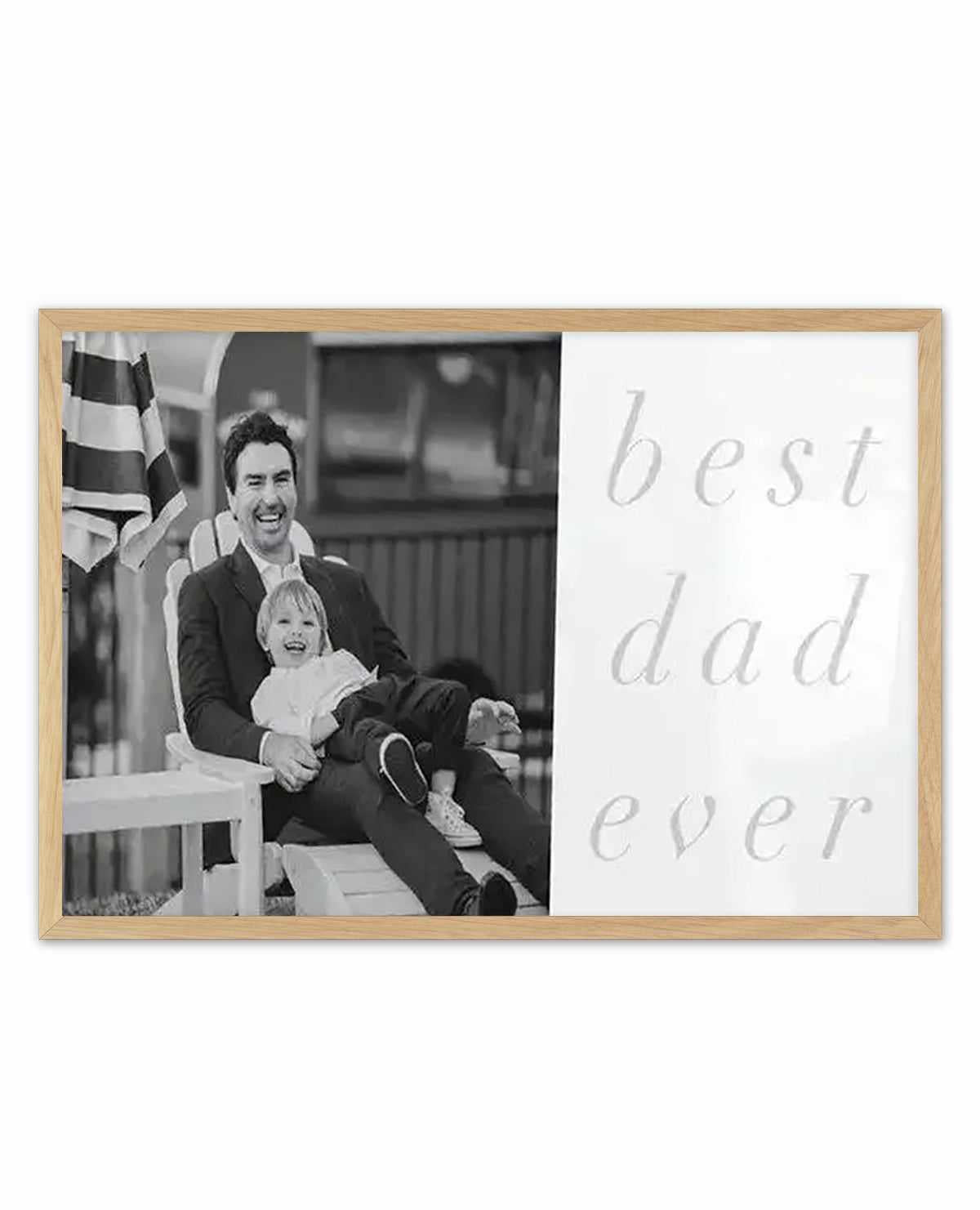 Black and white photograph printed on white background, framed in natural wood. Left side shows a smiling man in dark blazer holding a young child, both laughing, with striped flag visible behind them on a porch railing. Right side displays "best dad ever" in light gray cursive script. Horizontal landscape orientation, matte finish print with natural wood frame.
