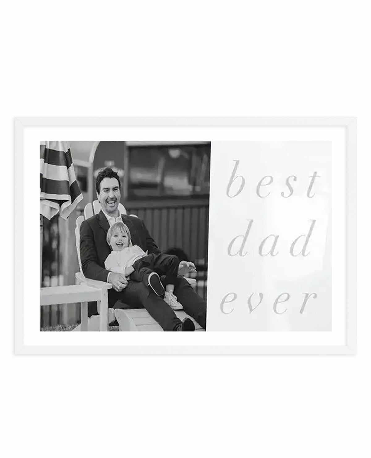 Black and white framed art print featuring a custom photo design. Left side displays a black and white photograph of a man and young child sitting together on a white bench with an American flag in the background. Right side shows light gray italic script text reading "best dad ever" on a white background. White frame with white border surrounding the entire composition.