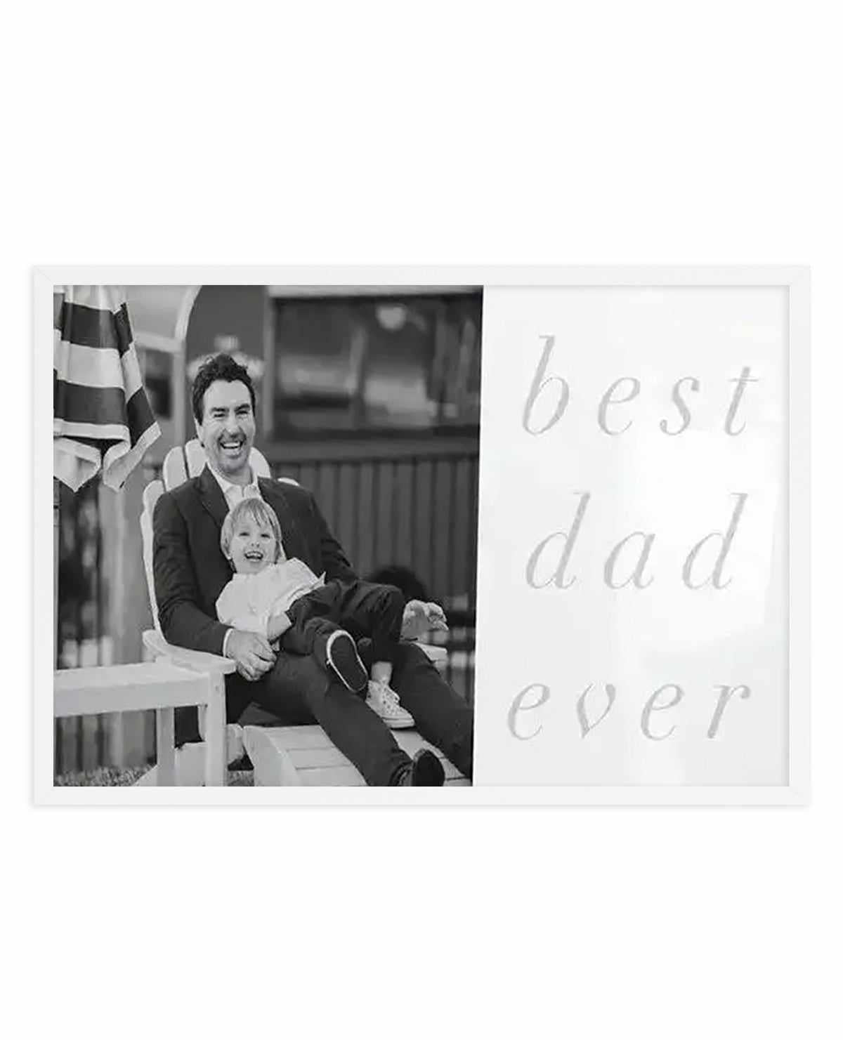 Black and white art print with white border featuring a custom photo on the left side showing a man and small child sitting together on white porch steps with a striped flag behind them, both smiling at camera. Right side displays text in light gray, serif font reading "best dad ever" arranged vertically. Print has matte finish and is mounted in white frame.