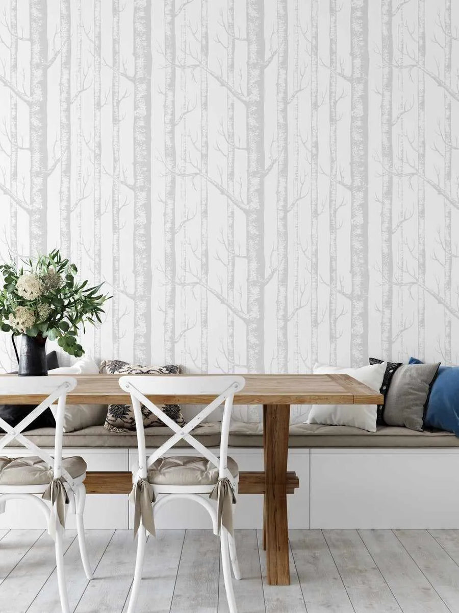 Lifestyle shot of a dining area with Birch Trees in Grey wallpaper. The wallpaper features a repeating pattern of tall, slender birch tree trunks in varying shades of light grey and white, with delicate bare branches extending outwards, creating a serene forest scene. The wallpaper covers the entire visible wall behind a light wooden dining table and a built-in white bench with neutral-toned cushions. Two white cross-back chairs with beige seat cushions and ties are visible in the foreground.