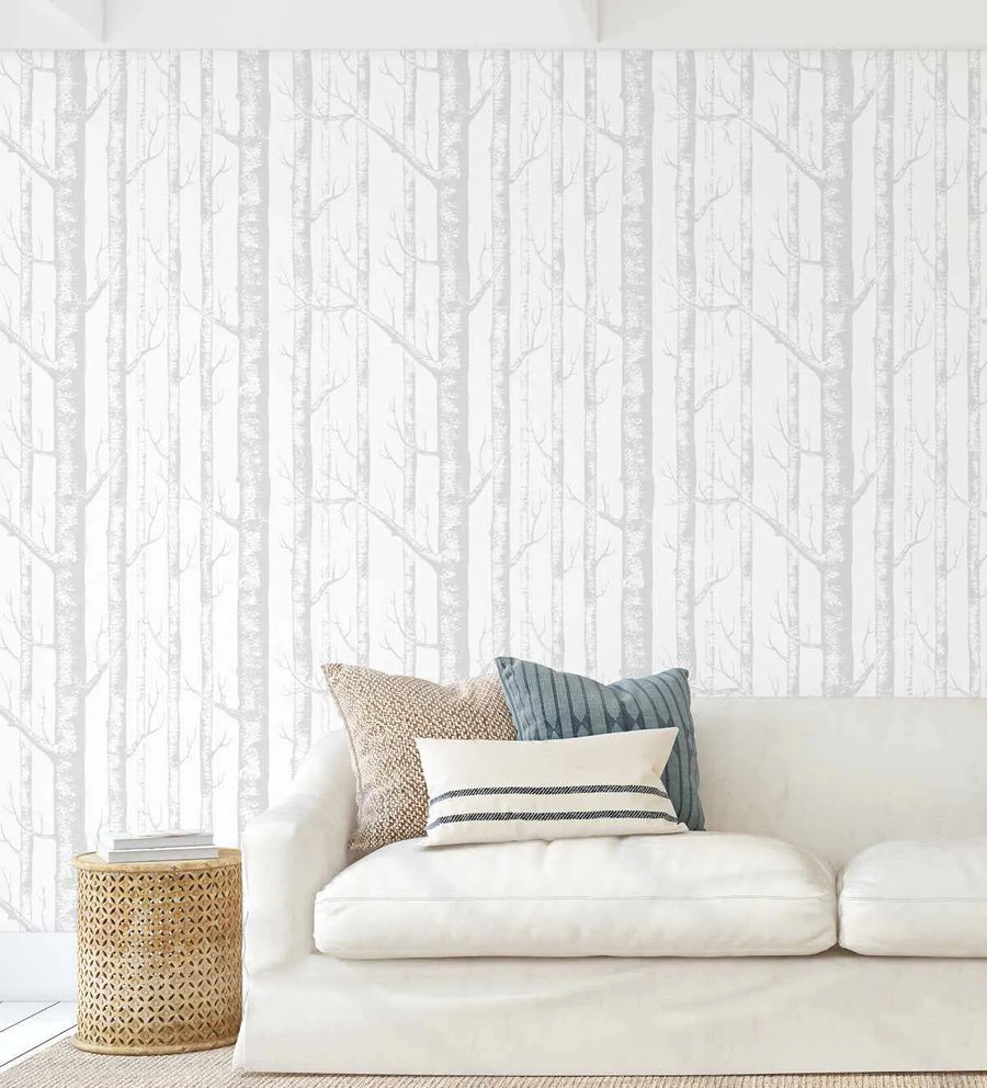 Lifestyle shot of a living room with Birch Trees in Grey wallpaper. The wallpaper features a repeating pattern of tall, slender birch tree trunks in varying shades of light grey against a clean white background, creating a serene forest scene. A white slipcover sofa with three decorative pillows (one textured beige, one solid blue, and one white with blue stripes) is positioned in front of the wallpaper. A round, light brown woven side table with books sits to the left of the sofa.