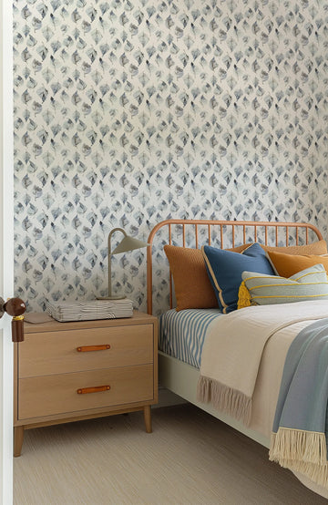 "Birch Leaves wallpaper featuring a repeating pattern of hand-painted birch leaves in muted blue-grey and charcoal on a soft white background. The botanical design has a loose, organic watercolour quality with visible brushstroke texture. The wallpaper is displayed on the wall behind a bed with a light wood headboard, a light wood bedside table with a lamp, and blue and brown pillows and blankets."