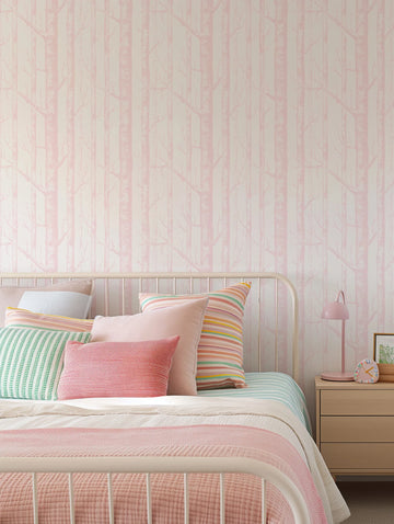 Lifestyle shot of a bedroom featuring wallpaper with a repeating pattern of slender birch trees in a soft pink hue against a light cream background. The wallpaper covers the wall behind a white metal bed frame, which is adorned with an assortment of pillows in shades of pink, coral, mint green, and multi-colored stripes. A light pink table lamp and a small, colorful clock sit on a light wood bedside table to the right.