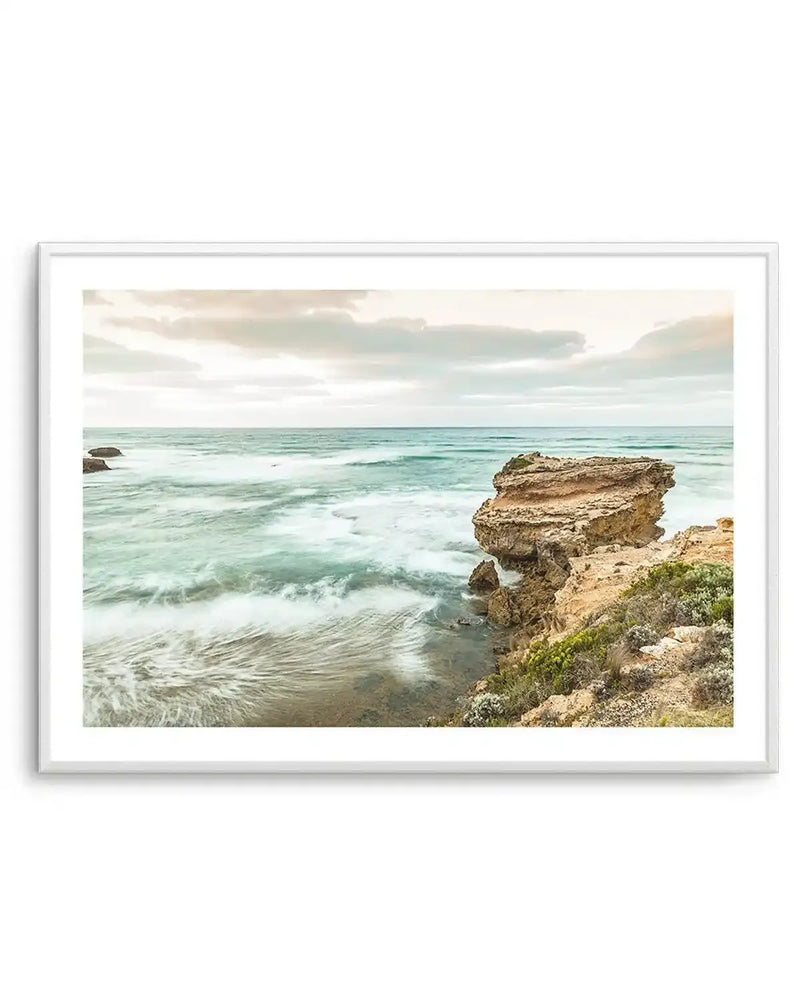 Framed photograph of a coastal scene with a white frame and white border. The composition features a wide expanse of ocean with gentle, long-exposure waves creating a soft, blurred effect, and a large, rugged rock formation on the right side of the frame. The sky is overcast with soft, muted clouds in pale grey and hints of peach. The water is a mix of turquoise and teal, and the foreground shows rocky shore with sparse green and brown vegetation.